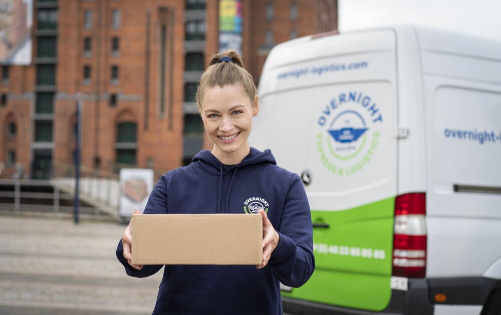 Eine Frau in einem blauen Hoodie lächelt und hält ein Pakete in den Händen, während ein Lieferwagen im Hintergrund steht.